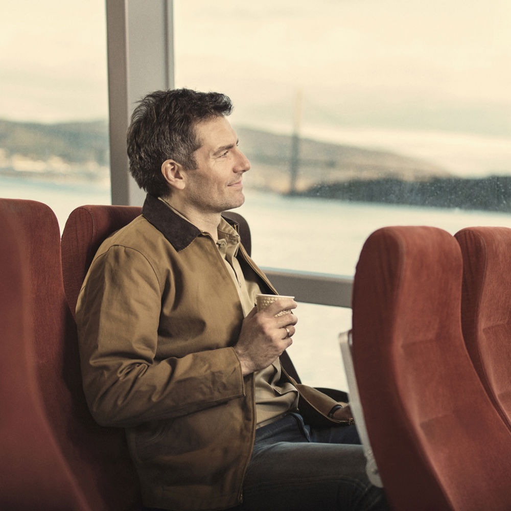 Mann med kaffikopp sitter i setet på en båt og ser ut av vinduet.