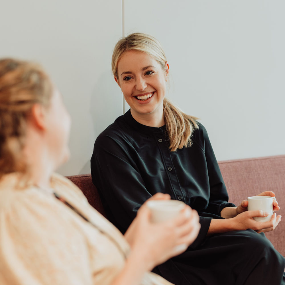 Bilde av Hanne i en sofa med en kaffekopp i hånden. Hun snakker med en annen kollega.
Foto: Skyss/Karoline Rage